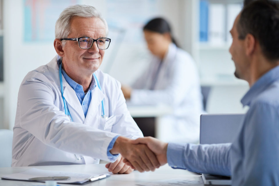 Smiling senior doctor shaking hands to his patient while they sitting at the table at office